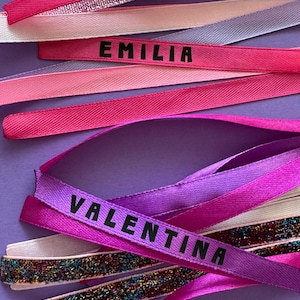 Può includere: Una collezione di nastri assortiti nelle tonalità del rosa, viola e argento, che include due nastri stampati con i nomi EMILIA e VALENTINA in lettere maiuscole nere. I nastri presentano texture varie, tra cui raso liscio e finiture glitterate.