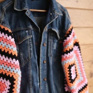 Könnte beinhalten: Blaue Jeansjacke mit bunten Häkelärmeln. Die Ärmel haben ein geometrisches Muster in Rosa, Orange, Schwarz und Blau. Die Jacke hat ein klassisches Knopfdesign und ist auf einem Holzbügel aufgehängt.
