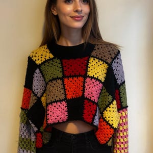Könnte beinhalten: Ein gecroppter Häkelpullover mit langen Ärmeln und einem Patchwork-Design. Der Körper des Pullovers ist schwarz mit bunten Quadraten in Rot, Gelb, Rosa, Grün und Lila. Die Ärmel sind eine Mischung aus den gleichen Farben. Der Pullover hat einen schwarzen Rundhalsausschnitt.
