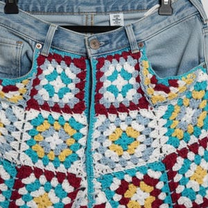 Op de afbeelding: Lichtblauwe spijkerbroek met haakdetails. Het haakontwerp heeft een kleurrijk patchworkpatroon met vierkanten in wit, blauwgroen, geel en bordeauxrood. De jeans hangt aan een zwarte hanger.