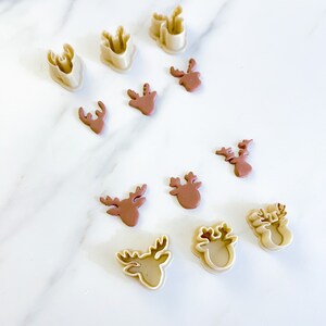 May include: A collection of beige plastic reindeer-shaped cookie cutters and corresponding brown clay reindeer cutouts. The set includes various sizes of deer head silhouettes with antlers, arranged in a scattered pattern across the frame.