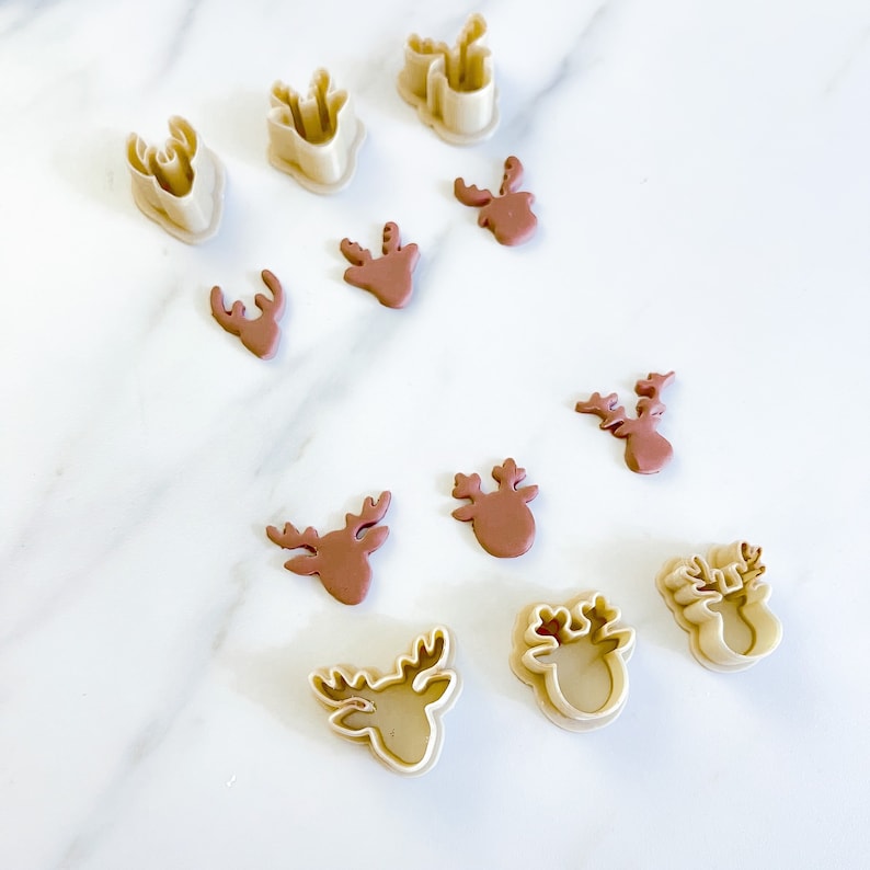 May include: A collection of beige plastic reindeer-shaped cookie cutters and corresponding brown clay reindeer cutouts. The set includes various sizes of deer head silhouettes with antlers, arranged in a scattered pattern across the frame.