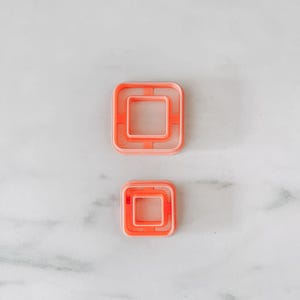 Peut inclure: Deux emporte-pièces carrés orange, l'un plus grand que l'autre, sont présentés sur un fond de marbre blanc. Les emporte-pièces ont une forme carrée dans un carré, créant un effet superposé. Ils sont probablement utilisés pour la pâtisserie.
