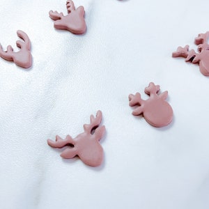 May include: Five small, flat, dusty rose-colored reindeer head shapes arranged in a scattered pattern. Each piece features a smooth, matte finish and clearly defined antlers, ears, and a rounded snout, crafted as individual decorative embellishments.