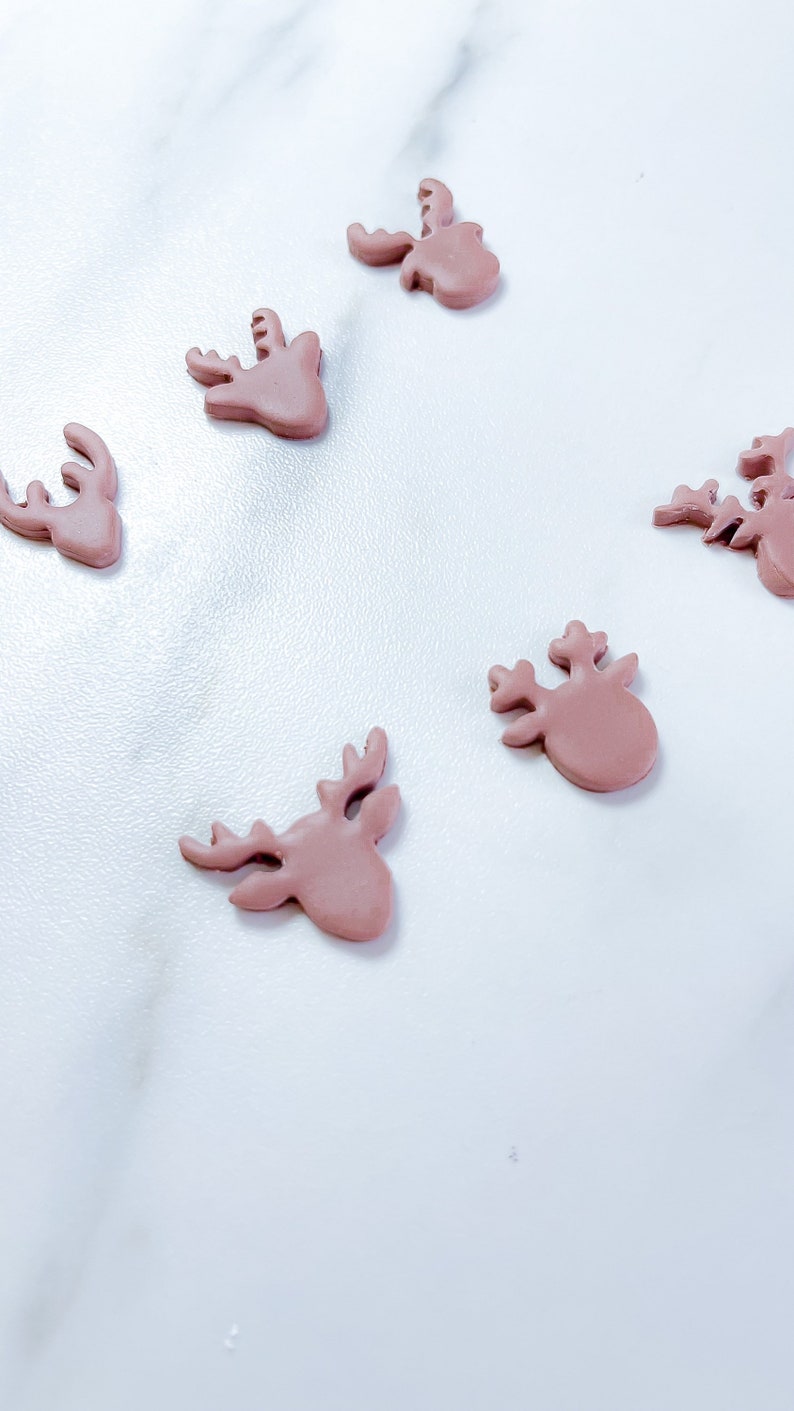 May include: Five small, flat, dusty rose-colored reindeer head shapes arranged in a scattered pattern. Each piece features a smooth, matte finish and clearly defined antlers, ears, and a rounded snout, crafted as individual decorative embellishments.