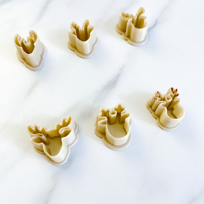 May include: Five beige plastic cookie cutters shaped like deer heads with antlers. Each cutter features a raised rim and a hollow center, designed to create outlines of deer silhouettes. The set includes various sizes and orientations of the deer head.