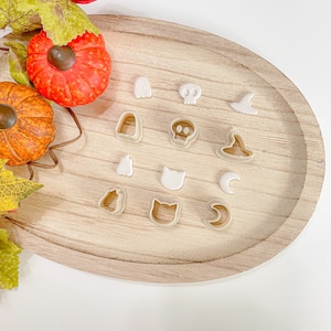 Peut inclure: Un ensemble de 12 emporte-pièces sur le thème d'Halloween sur une surface en bois. Les emporte-pièces sont en forme de fantôme, de crâne, de chapeau de sorcière, de chat, de croissant de lune et de citrouille.