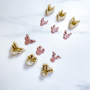 May include: A collection of beige plastic reindeer-shaped cookie cutters arranged alongside several small, light brown reindeer-shaped cutouts made from dough or clay, showcasing the detailed antler and head silhouettes of the animal figures.