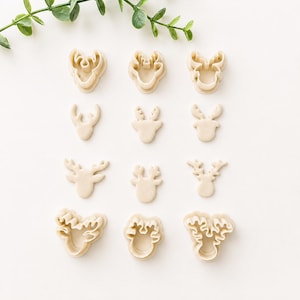 May include: A collection of twelve beige cookie cutters arranged in four rows, featuring various reindeer head shapes with distinct antler designs. The set includes both outline cutters and detailed impression cutters in a light cream color.