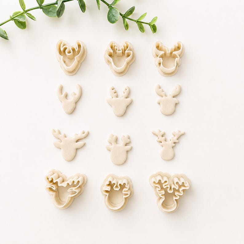 May include: A collection of twelve beige cookie cutters arranged in four rows, featuring various reindeer head shapes with distinct antler designs. The set includes both outline cutters and detailed impression cutters in a light cream color.