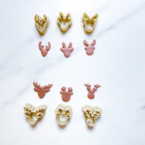 May include: A collection of six beige plastic reindeer-shaped cookie cutters and six corresponding brown reindeer-shaped cutouts arranged in rows. The cutters feature detailed antler designs, while the cutouts show the solid reindeer head shapes.