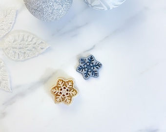 Puede incluir: Dos adornos con forma de copo de nieve, uno es de color azul claro y el otro es de color beige claro. Los adornos están sobre una superficie de mármol blanco.