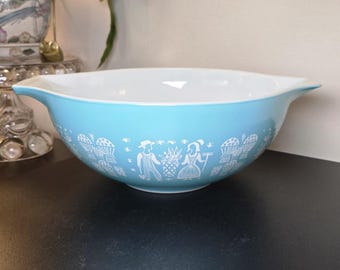 Op de afbeelding: Een lichtblauwe mengkom met een witte binnenkant en twee handvatten aan de zijkanten. De buitenkant is voorzien van een herhalende witte grafische print van figuren met hoeden en schorten naast manden met producten en korenaren.