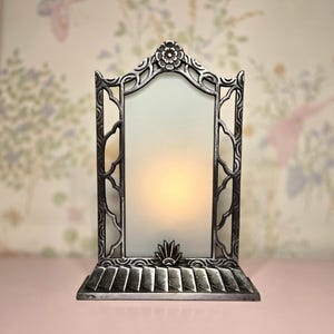 May include: A decorative silver-toned candle holder with an ornate, Art Nouveau-inspired design. The holder features a rectangular frosted glass panel, a floral top, and a stylized base. The candle inside emits a warm, inviting glow.