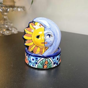 May include: A ceramic trinket box featuring a lid with a split sun and moon face design in yellow, blue, and brown. The base is cylindrical with a hand-painted pattern of blue, green, and orange floral and geometric motifs on a white background.