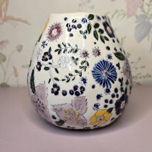 May include: A white ceramic vase with a rounded shape, adorned with a colorful floral pattern. The design includes blue, purple, yellow, and green flowers and leaves. The vase has a glossy finish and sits on a pink surface.