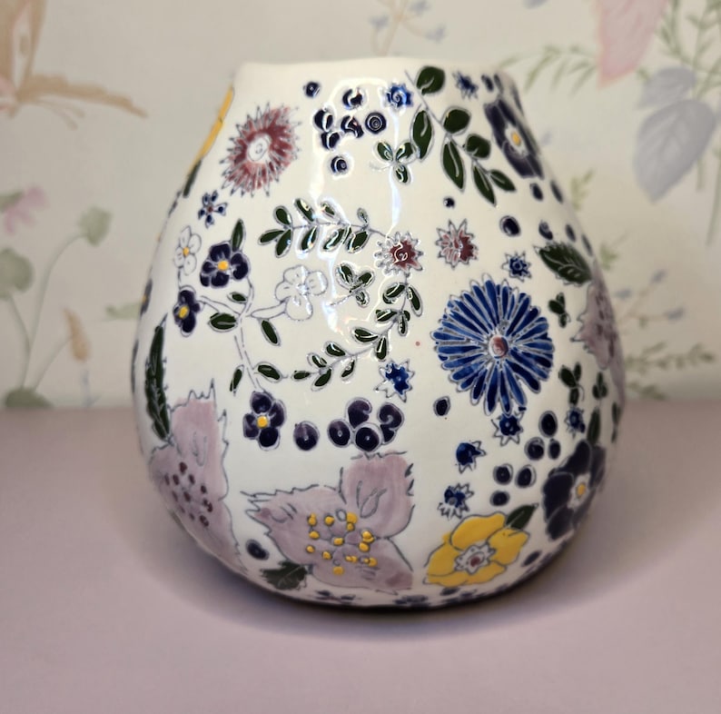 May include: A white ceramic vase with a rounded shape, adorned with a colorful floral pattern. The design includes blue, purple, yellow, and green flowers and leaves. The vase has a glossy finish and sits on a pink surface.