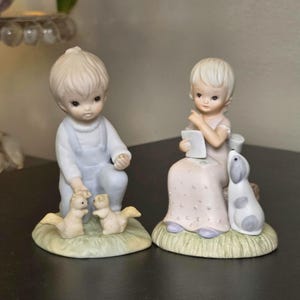 May include: Two porcelain figurines featuring children with blonde hair. One child wears light blue overalls and kneels beside two small squirrels. The other child wears a pale pink dress, sits on a base, and holds a white paper near a small dog.