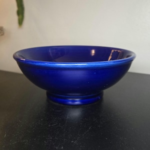 May include: A deep cobalt blue ceramic bowl with a smooth, glossy finish. The bowl features a wide, open circular rim that tapers down to a smaller, raised pedestal base, creating a distinct silhouette with a uniform, solid color throughout.