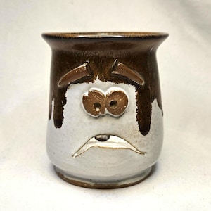 May include: A handmade ceramic mug with a sad face design. The mug features a white base with brown accents and a brown rim. The face has brown eyebrows, eyes, and a downturned mouth, creating a distressed expression. The mug is suitable for beverages.