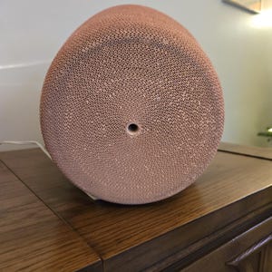 May include: A light brown, egg-shaped object with a textured surface made up of small, evenly spaced holes. The object has a single, central hole on one end. It rests on a dark wooden surface.