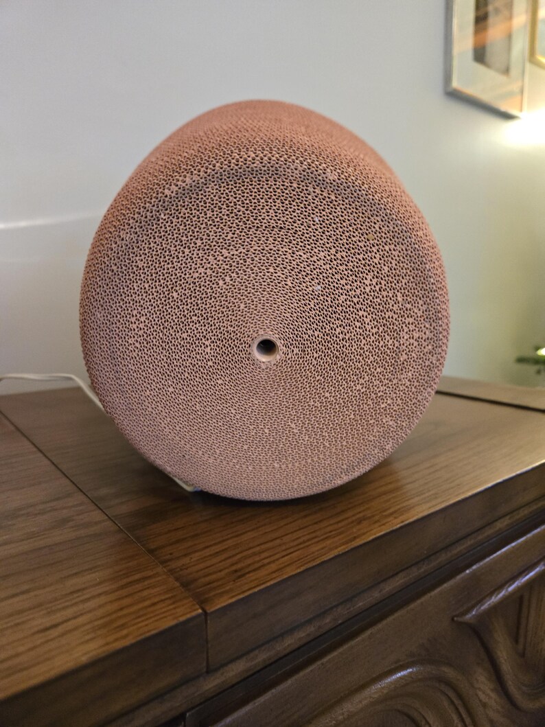 May include: A light brown, egg-shaped object with a textured surface made up of small, evenly spaced holes. The object has a single, central hole on one end. It rests on a dark wooden surface.