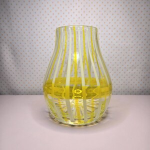 May include: A clear glass vase with vertical yellow and white stripes. The vase has a rounded body and a flared top. The stripes are evenly spaced and create a visually appealing pattern. The vase is sitting on a light pink surface.