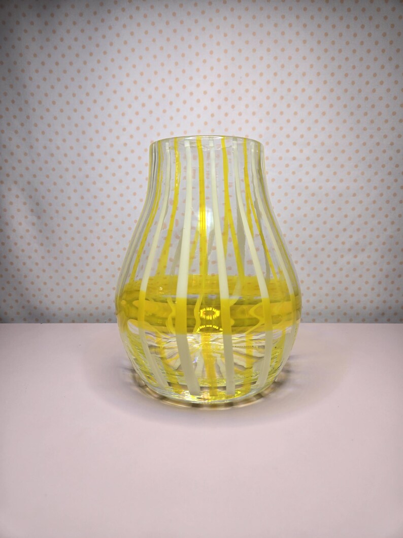 May include: A clear glass vase with vertical yellow and white stripes. The vase has a rounded body and a flared top. The stripes are evenly spaced and create a visually appealing pattern. The vase is sitting on a light pink surface.
