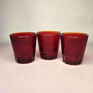 May include: Three ruby red drinking glasses. The glasses have a slightly tapered shape with a decorative pattern near the base. The rims of the glasses have a golden hue. The glasses are made of glass.