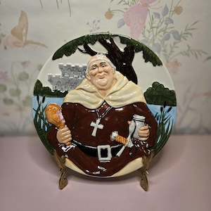 May include: A decorative ceramic plate featuring a jovial monk holding a chicken leg and a jar. The plate has a brown, white, and beige color scheme with a castle and tree design in the background. The plate is displayed on a gold-colored stand.
