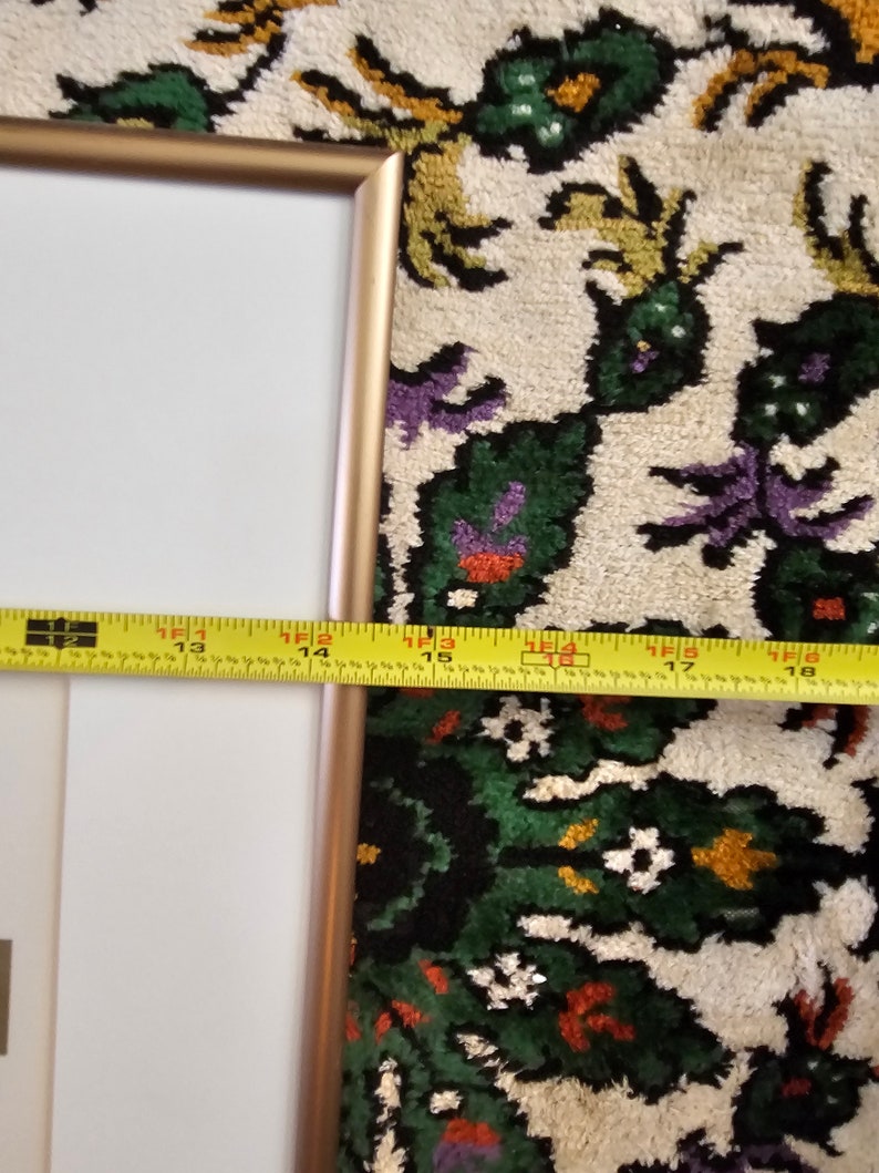 May include: A gold-coloured picture frame with a white mount and a white background. The frame is sitting on a colourful rug with a floral pattern.