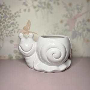 May include: A white ceramic snail-shaped planter with a smiling face and a spiraled shell. The planter sits on a pink surface, with a floral wallpaper background. The snail planter is a decorative item.