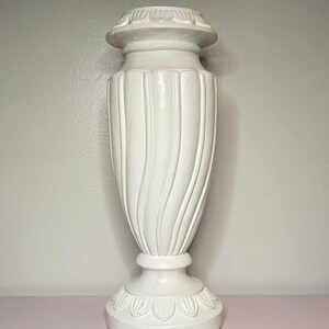 May include: A white, decorative urn with a fluted, curved design. The urn has a round base and a detailed top. The overall height is approximately 18 inches.