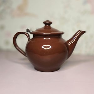 May include: A brown ceramic teapot with a lid, handle, and spout. The teapot has a glossy finish and a rounded body. The handle is curved, and the spout is slightly angled. The lid has a small knob on top.