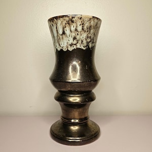 May include: A ceramic vase with a flared rim and a pedestal base. The upper portion features a white glaze with brown drip patterns, while the lower body and base are finished in a solid, metallic dark brown glaze with a smooth, reflective surface.