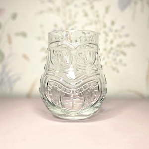 May include: Clear glass tiki-style tumbler with a detailed carved face design. The glass features a wide, smiling mouth, large eyes, and a textured surface. This unique drinking glass is perfect for tropical cocktails.