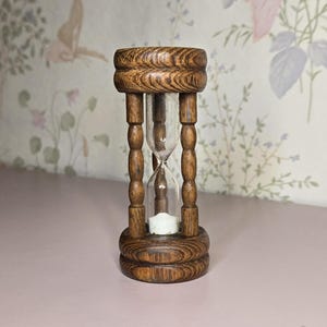 May include: A wooden hourglass with a clear glass center. The hourglass is made of dark brown wood and has a decorative, carved design. The sand timer is set against a floral wallpaper background.