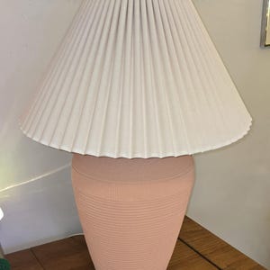 May include: A peach-coloured table lamp with a pleated off-white lampshade. The lamp base has a textured, ribbed design. The lamp is on a wooden table.