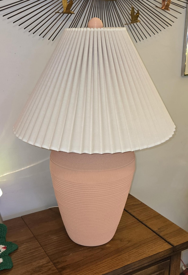 May include: A peach-coloured table lamp with a pleated off-white lampshade. The lamp base has a textured, ribbed design. The lamp is on a wooden table.