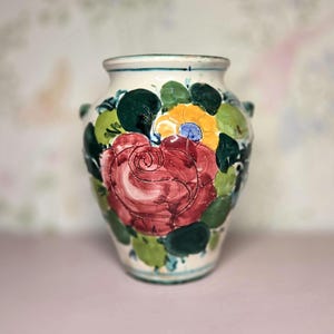 May include: A hand-painted ceramic vase with a cream-colored base. The vase features a large red rose with yellow, green, and blue floral accents. Two small handles are on the sides. The rim and base have a thin blue stripe.