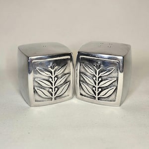 May include: A pair of silver-colored salt and pepper shakers. Each shaker is a cube shape with rounded edges. A raised leaf design is centered on the front of each shaker. The top of one shaker has three holes, and the other has one hole.