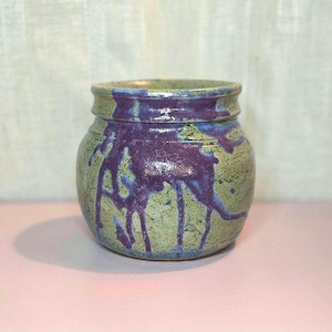 May include: A handmade ceramic jar with a rounded body and a slightly flared rim. The jar is primarily green with blue accents and striking purple drips. The jar is likely used for storage or decoration.