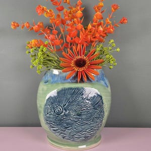 May include: A ceramic vase with a fish design, filled with orange and green flowers. The vase is primarily green with blue accents and a detailed, textured fish illustration. The flowers add a vibrant contrast to the vase's colors.