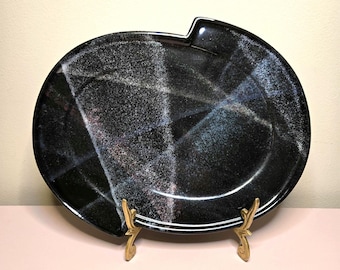May include: An oval-shaped black ceramic platter featuring a glossy finish with abstract white and blue speckled cross-hatch patterns. The platter has a distinct notched edge at the top and is displayed on a gold-toned metal stand.