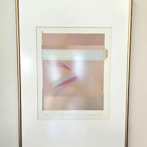 May include: A framed abstract art print with a pale pink, white, and grey colour scheme. The print features geometric shapes and lines, with a subtle texture. The artist's signature and date are visible in the lower right corner.