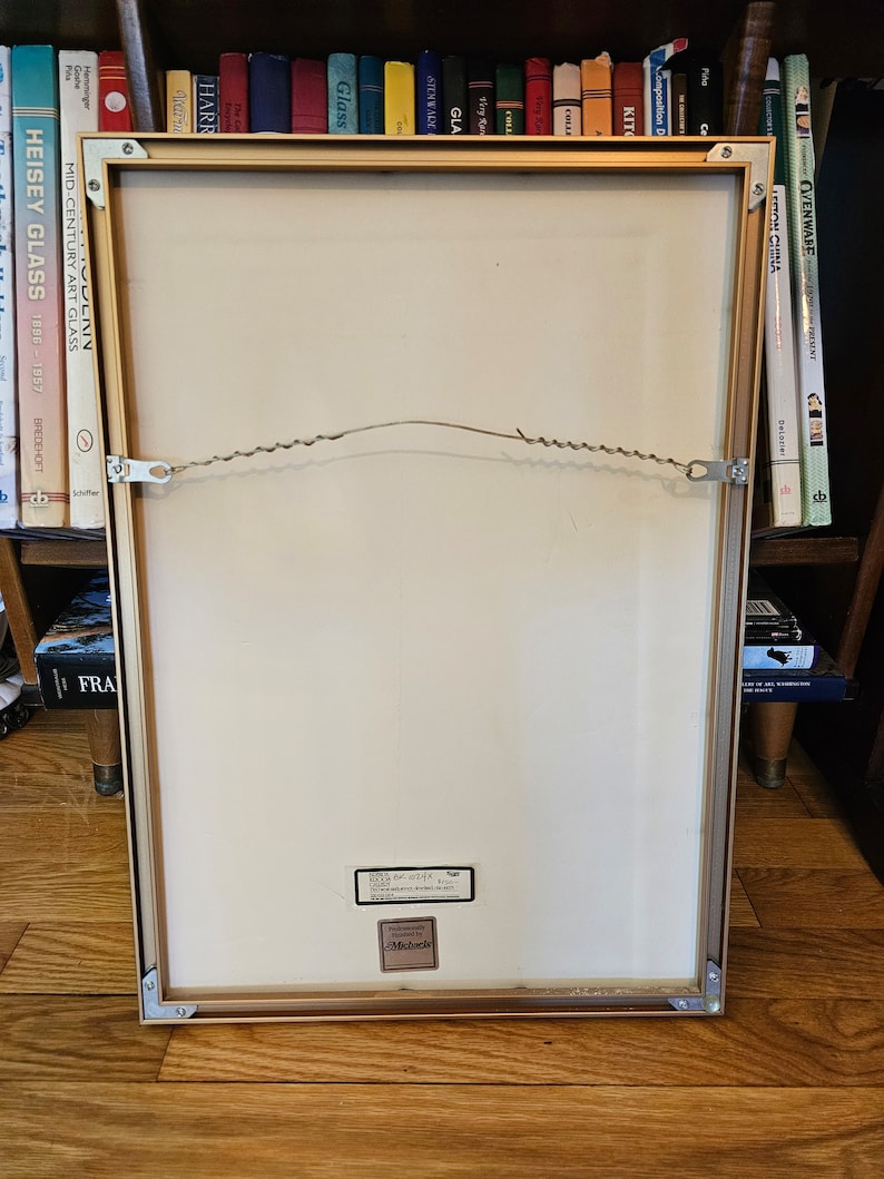 May include: A gold-coloured picture frame with a white mount and a white background. The frame has a wire hanger on the back and a sticker that says "Professionally Framed by Michaels".