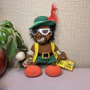 May include: A plush doll of a dark-skinned character wearing a green hat with a red feather, white-rimmed glasses, and orange shoes. The doll has a yellow vest, green shorts, and a black belt. The tag reads "Trash Talkers".