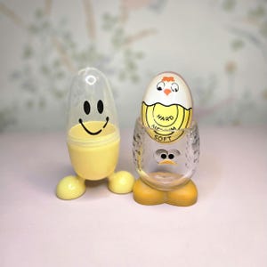 May include: Two novelty egg timers. One is yellow with a smiley face and clear top. The other is a clear egg cup with a chick design, holding an egg with "Hard, Medium, Soft" text.