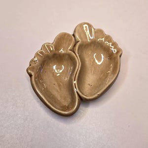 May include: A ceramic dish in the shape of two baby footprints. The dish is a light brown color with a glossy finish. The footprints are detailed with toe and arch impressions. A decorative item for a nursery or baby shower.