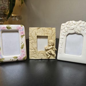 May include: Three rectangular picture frames. The left frame is white with pink flowers and green leaves. The center frame is beige with a textured surface and a small cherub figure. The right frame is white with a raised leaf and fruit pattern.